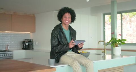 African American man sitting on kitchen island smiling while holding tablet in modern kitchen