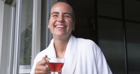 Smiling woman in white bathrobe holding glass teacup enjoying serene morning by window