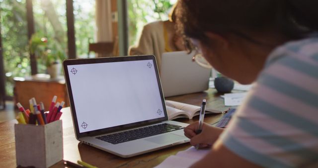 Young student focusing on homework at desk with open laptop. Ideal for educational content, online learning resources, or study tips.