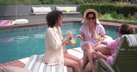 Friends sipping cocktails poolside laughing in sun hats and sunglasses backyard summer