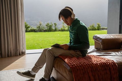 Asian man relaxing with headphones and tablet on leather sofa overlooking green garden