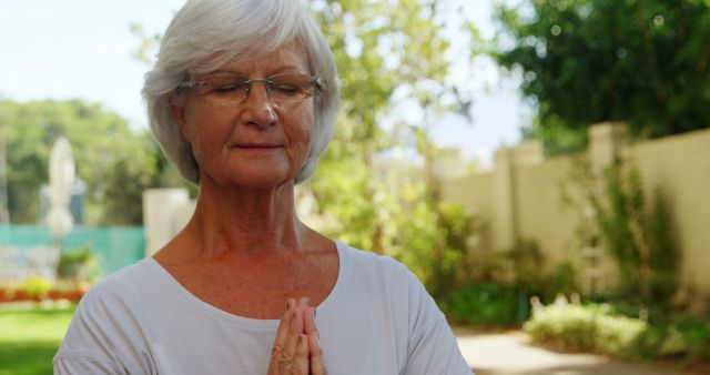 A senior Caucasian woman is practicing meditation outdoors, with her eyes closed and hands in a prayer position, with copy space. Her serene expression suggests a moment of peace and mindfulness in a tranquil garden setting.