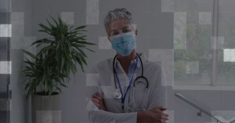 Senior clinician standing arms crossed wearing mask and stethoscope in clinic hallway