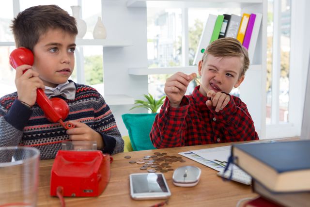 Two children role-playing as business executives, using a red phone and counting coins at a desk. Great for concepts of childhood imagination, education, learning through play, and aspiring young professionals.