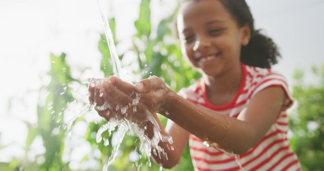 This stock photo features a smiling African American girl washing her hands with water in an outdoor natural setting. The vibrant greenery provides a fresh and clean background. These themes make it perfect for use in articles or marketing promoting hygiene and health benefits, environmental conservation, or showcasing joyful childhood moments during summer.