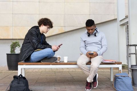 Diverse teen students sitting on campus bench using smartphones with coffee and backpacks