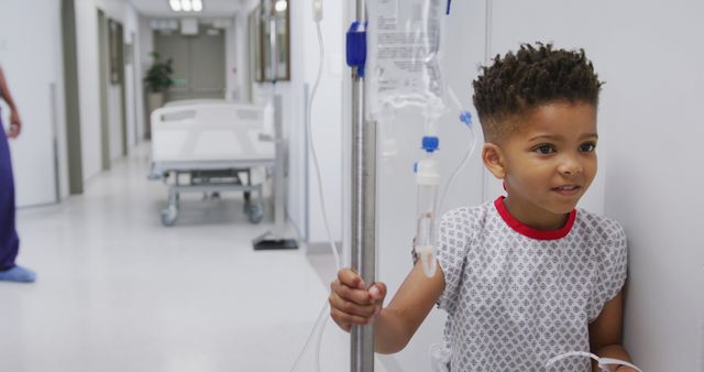 Young African American boy standing in hospital corridor, holding an IV drip and smiling. Can be used for healthcare, pediatric care, medical treatment, and hospital environment concepts.