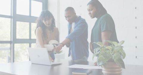 Diverse team collaborating around laptop in bright modern office, man pointing and guiding