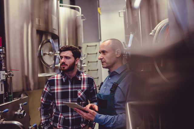Brewery owner and worker inspecting machinery in a brewery. Ideal for use in articles about beer production, industrial processes, teamwork in manufacturing, and quality control in brewing.
