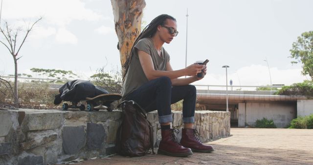 Young man with long dreadlocks and sunglasses using smartphone, enjoying a moment outdoors. He is seated on a stone bench with a skateboard and a bag beside him. Useful for themes such as technology, urban lifestyle, youth culture, fashion trends, and relaxation.