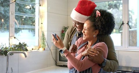 Couple hugging in festive kitchen wearing santa hat and aprons while looking at smartphone