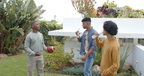 Multicultural young men chatting and drinking bottles on backyard lawn with football