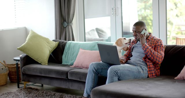 Man telecommuting from his living room, using laptop while on phone call, dog nearby on sofa. Perfect for articles about remote work, achieving work-life balance, productivity at home, and the impact of pets on mental health.