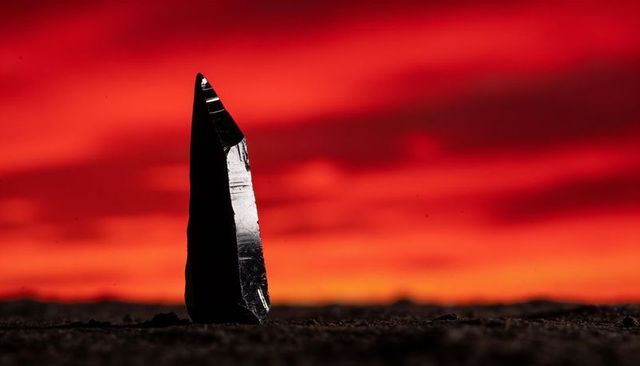 Obsidian monolith rising from granular ground against fiery red-orange sunset
