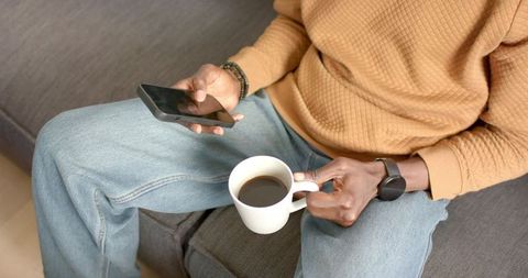 African American man sitting on sofa holding smartphone and coffee mug in casual homewear