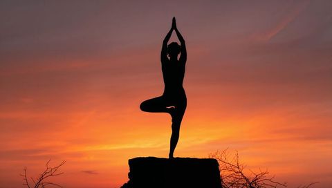 Silhouetted woman practicing tree pose on cliff at vibrant orange and purple sunset