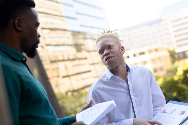 African american mid adult businessman talking over data to albino male colleague in office balcony. unaltered, abnormal, data, corporate business, teamwork, meeting, occupation and office concept.