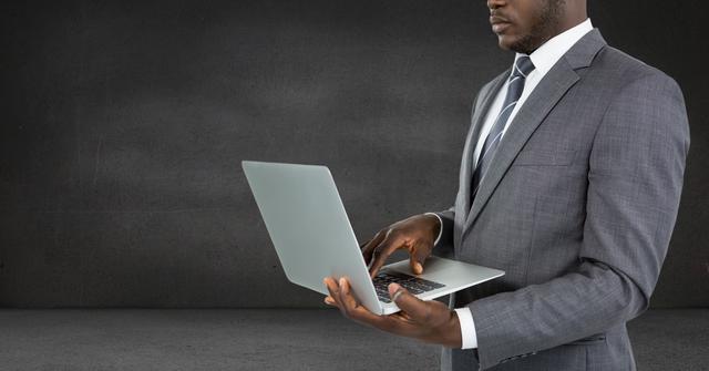 Businessman wearing formal suit using laptop against grey background. Ideal for illustrating concepts of corporate work, business technology, professional attitude, job ads, company websites, modern office environments, and remote work solutions.