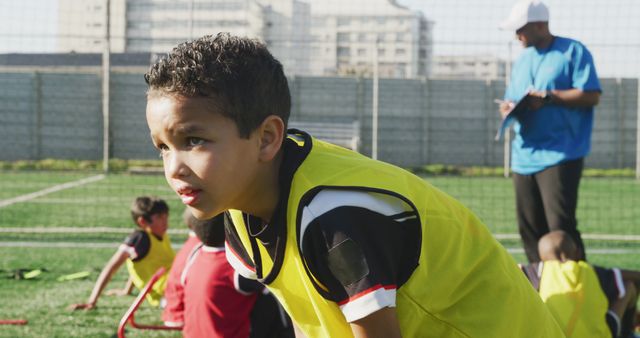 Young boy intensely focusing during soccer training on a green field, with his coach instructing in the background. Ideal for use in articles about youth sports, teamwork, physical activities for children, and outdoor exercises.