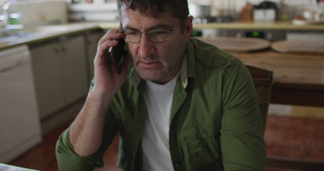 Middle-aged man in casual clothing talking on phone in kitchen. Useful for depicting attention to conversation or problem solving at home, discussing important matters, modern communication, and everyday life scenarios.
