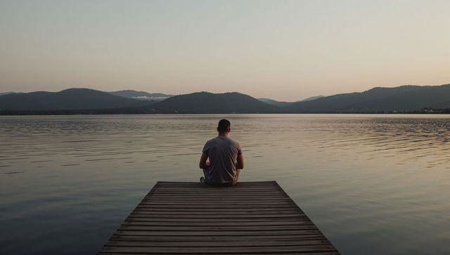 Solitary man sitting on wooden dock facing calm lake at sunrise with mountain horizon