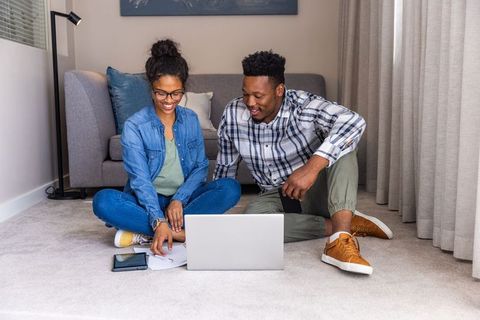 Young couple working from home on laptop while sitting cross-legged on living room floor