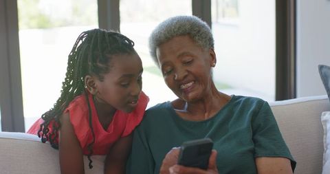 African grandmother and granddaughter sharing smartphone on sofa teaching digital skills