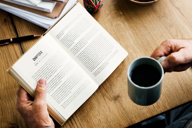 Person holding open book and drinking coffee on wooden table. Ideal for blogs on education, study tips, relaxing routines, book clubs, and lifestyle articles.