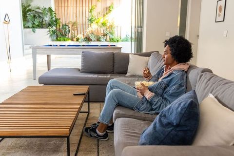 African american woman relaxing on couch snacking popcorn watching tv in modern living room