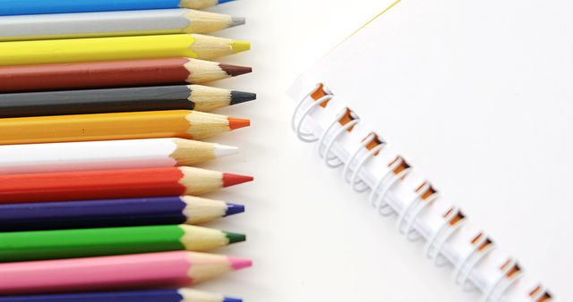 Colored pencils are neatly aligned on a white surface, pointing towards a blank spiral notebook, with copy space. Art supplies like these are often used by students, artists, and creative professionals to sketch and brainstorm ideas.