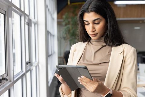 Indian businesswoman using tablet by office windows wearing beige blazer and smartwatch