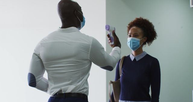 A male staff member is conducting a temperature check with an infrared thermometer for a female employee at the entrance of an office. Both are wearing protective face masks, emphasizing health and safety protocols during the COVID-19 pandemic. This image is perfect for topics related to workplace safety, health precautions, pandemic measures, office environments, and employee well-being.