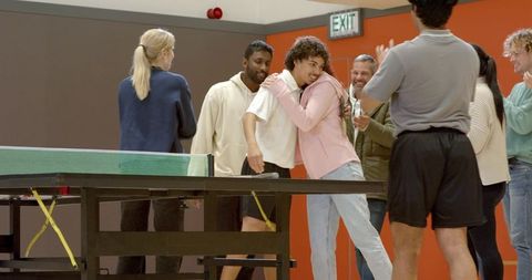 Diverse team hugging and cheering around table tennis in recreation room