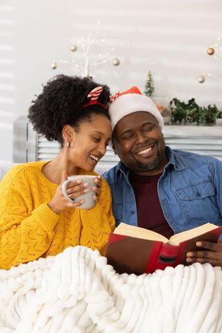African american couple cuddling on sofa under blanket holding mug and reading book