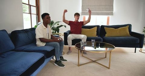 Young friends cheering while playing video games on blue velvet sofas in living room