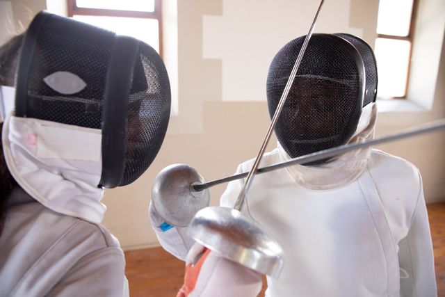 Two people in a fencing duel, wearing protective fencing clothes, holding sabers, in a gym. Sport and working out.
