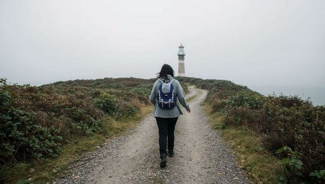 Hiker walking toward lighthouse on foggy coastal trail with backpack and boots