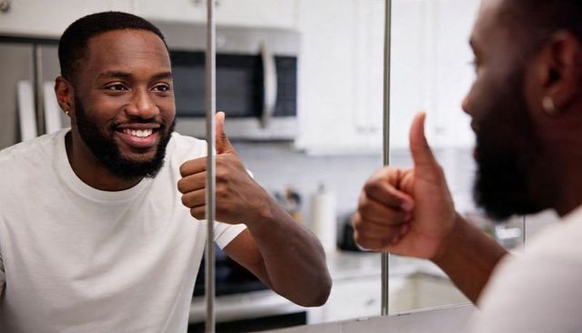 Smiling black man giving thumbs-up at mirror in modern kitchen, showing self confidence