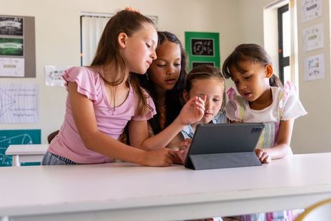 Diverse elementary girls collaborating on tablet, leaning and pointing in bright classroom