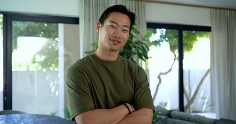 Confident asian man standing with arms crossed in modern living room wearing olive tee