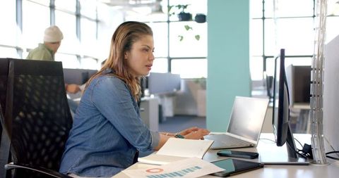 Mid adult woman working at desk with laptop and monitor in modern open-plan office