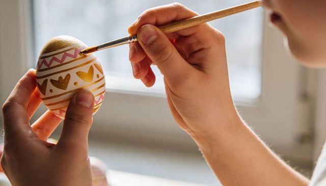 Child painting decorative easter egg with gold and pink patterns near window light