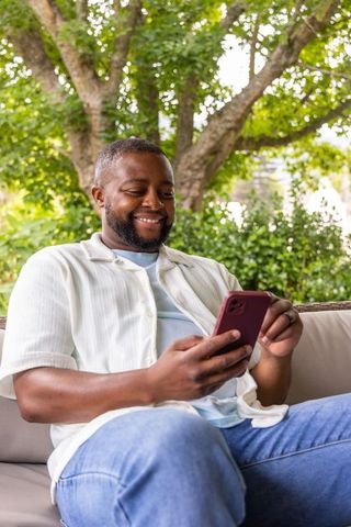 African american man smiling while using smartphone on outdoor sofa under leafy tree