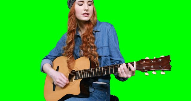Young red-haired woman sitting and playing an acoustic guitar against a green screen background. Wearing a denim shirt and a headband, she looks focused on her music. Ideal for use in music videos, educational content about guitar playing, music instruction, or promotional videos. The green screen allows for easy background replacement.