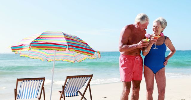 Senior couple wearing swimsuits standing on beach, holding cocktails and smiling. Colorful umbrella and two empty chairs in background. Perfect for travel advertisements, retirement concepts, and lifestyle blog visuals.