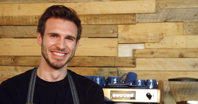 Cheerful male barista smiling warmly, standing in cafe with modern espresso machine and rustic wooden wall behind him. Ideal for websites, blogs, advertisements promoting coffee shops, barista training, or positive customer service experiences.