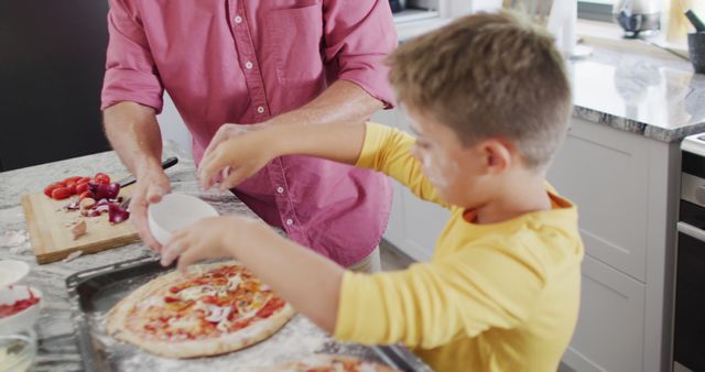 Father and son are enjoying a culinary bonding experience by making homemade pizza in the kitchen. The son is sprinkling cheese while the father provides guidance, showcasing a special moment full of fun and learning. Perfect for use in blogs, articles, or advertisements related to family activities, cooking, parenting, and child development.