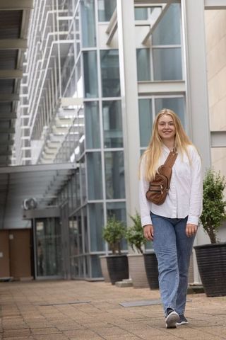 Smiling woman walking past modern glass office building wearing white shirt and brown sling bag