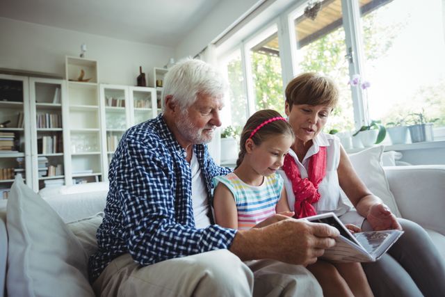 Grandparents and granddaughter sitting on couch in living room, looking at photo album together. Ideal for use in family-oriented content, advertisements about family bonding, or articles on intergenerational relationships and preserving memories.