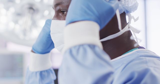 Image of african american male surgeon tying face mask in operating theatre, copy space. Hospital, medical and healthcare services.
